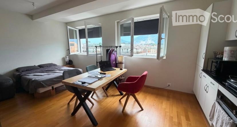 A modern, well-lit bedroom with a large window offering a scenic view. The room features a bed with dark bedding, a wooden desk with a laptop, a pink chair, and a small kitchen area with cabinets and a microwave.