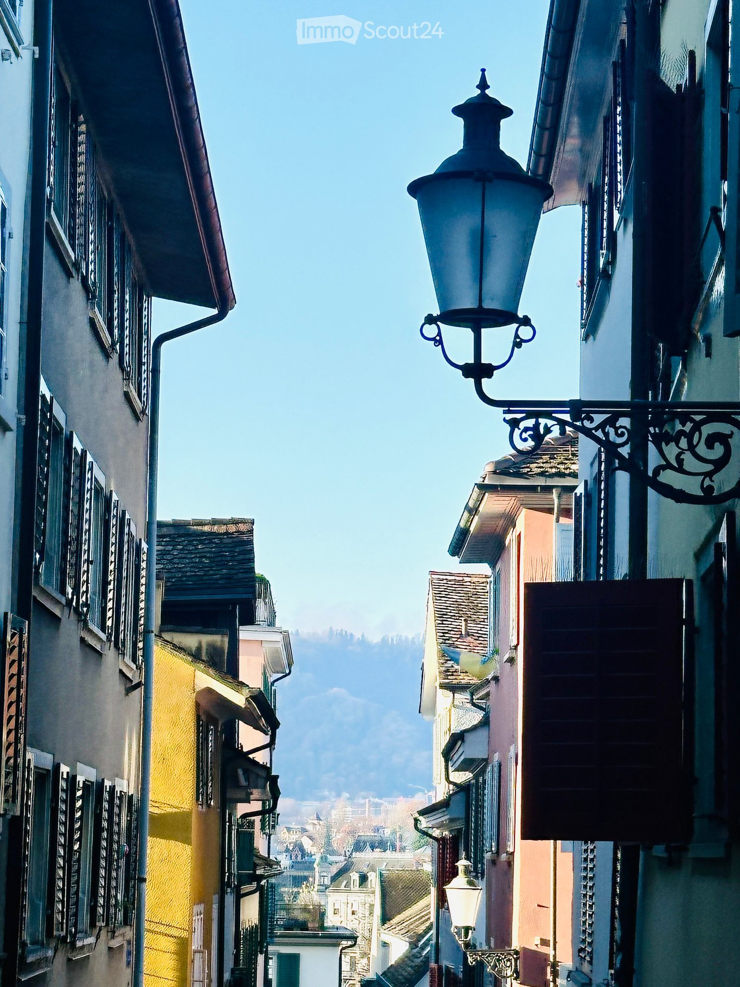 A narrow street lined with colorful buildings, featuring ornate street lamps and a view of distant rooftops and mountains.