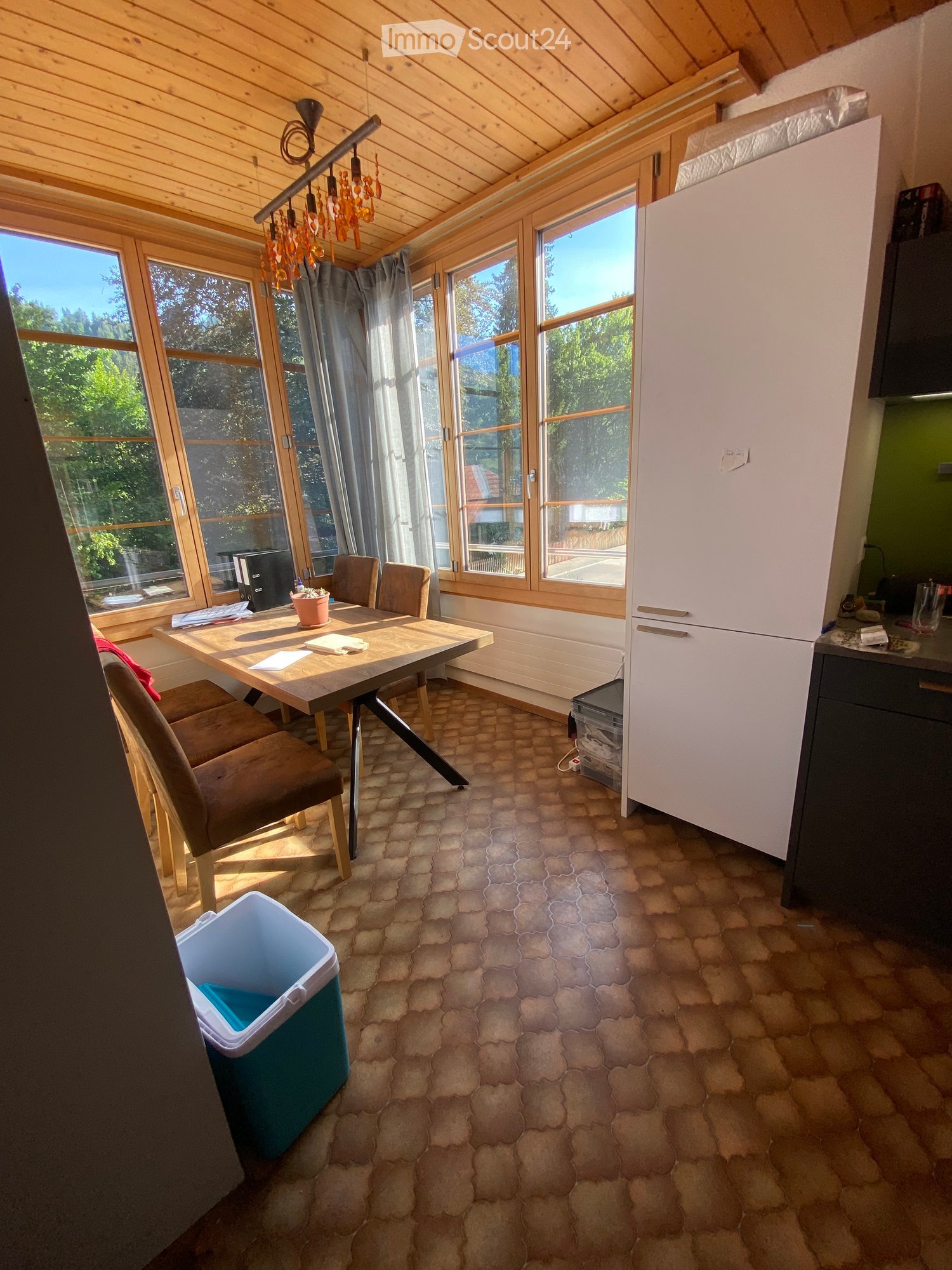 Bright room with wooden ceiling, large windows, a dining table with chairs, a refrigerator, and a trash bin.