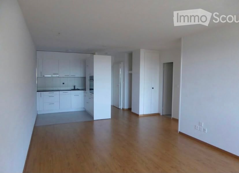 Bright, open-plan living and kitchen area with white cabinets, a sink, and a refrigerator. The floor is wooden, and the walls are white.