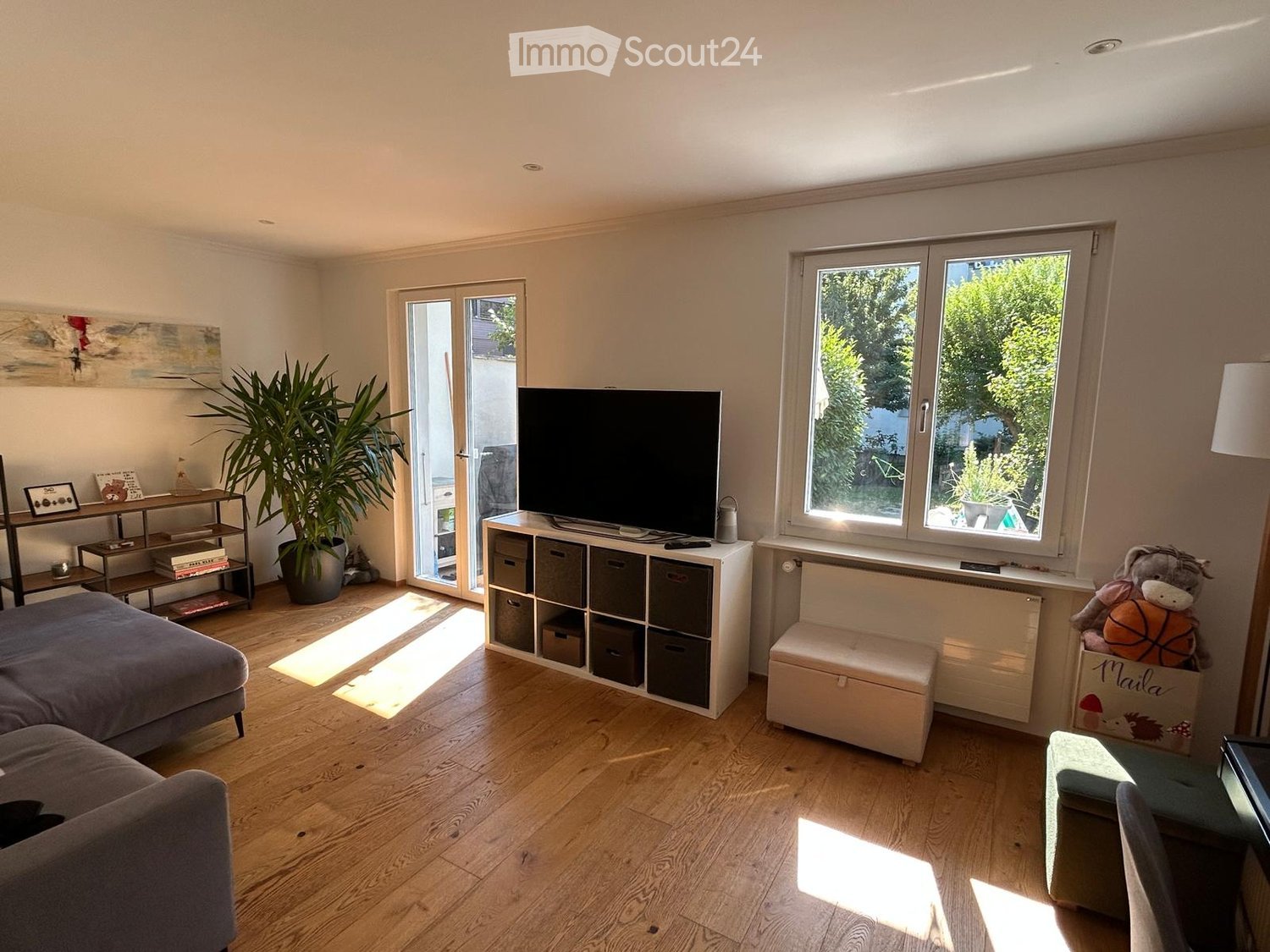 Bright living room with wooden flooring, large windows, and a sliding glass door leading to a balcony. Features a grey sectional sofa, a white storage unit with multiple compartments, a small round table, a potted plant, and a decorative stool.