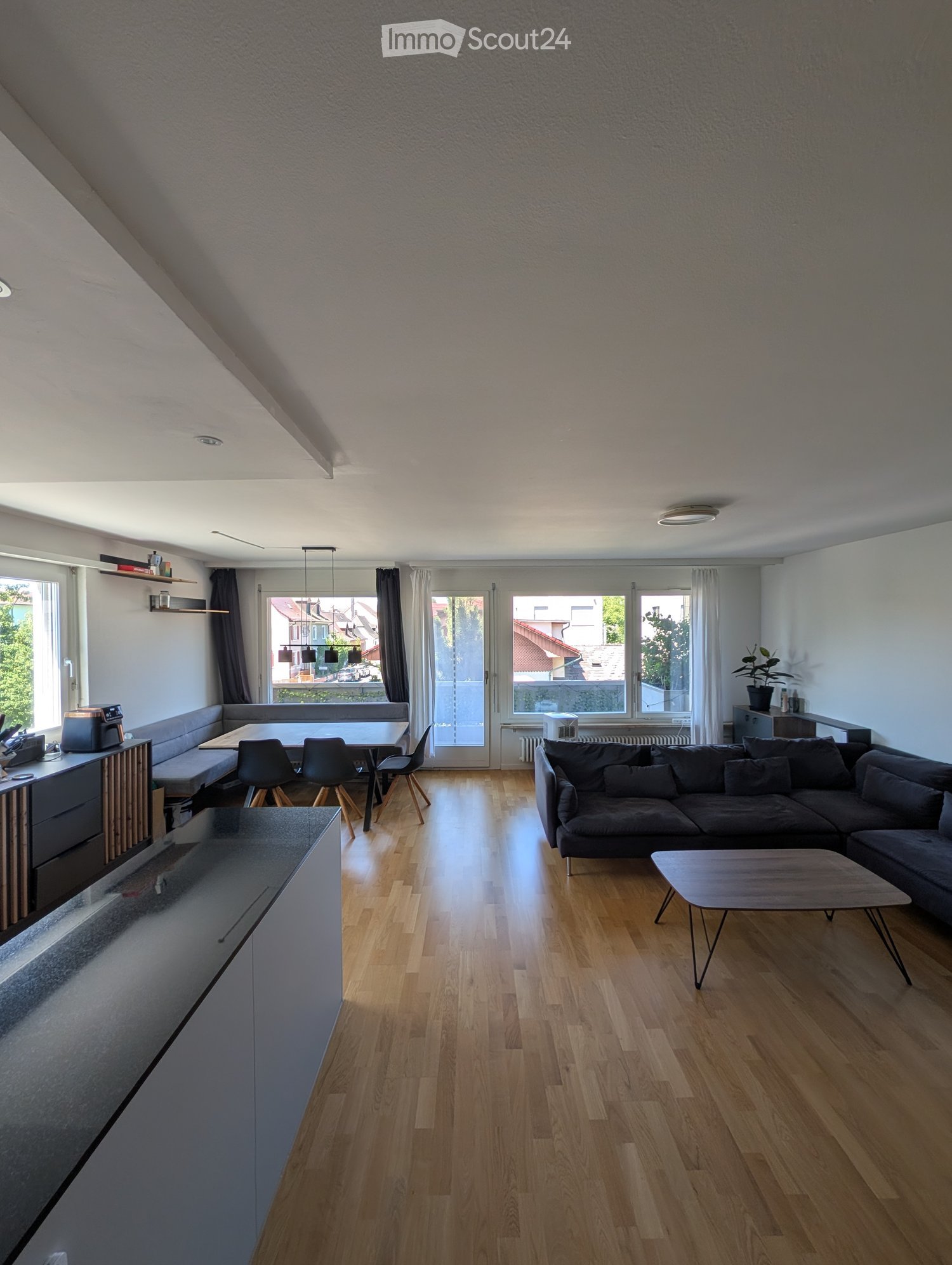 Modern living room with wooden flooring, large windows, a sectional sofa, coffee table, dining area with chairs, and a small kitchen counter.