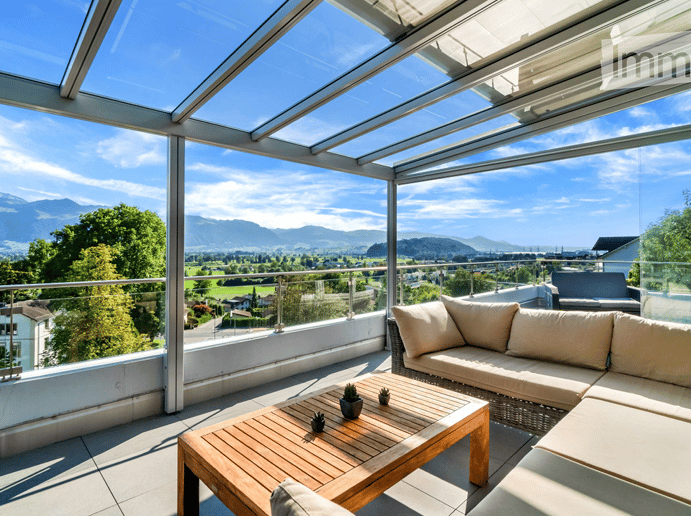 A modern glass-enclosed balcony with a panoramic mountain view, featuring a wooden coffee table, a cream-colored sofa, and small potted plants.