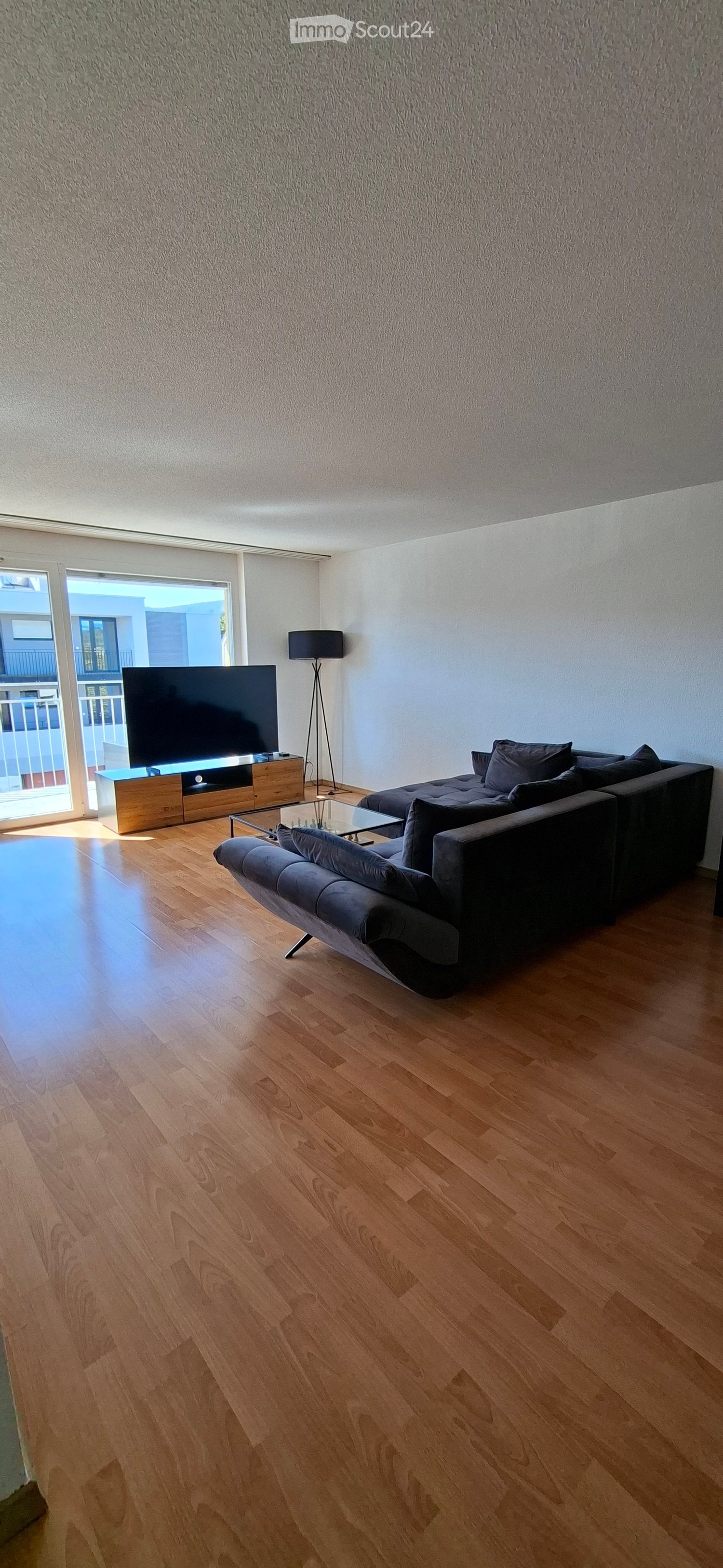 Modern living room with wooden flooring, a sectional sofa, a coffee table, a television, and a floor lamp near a large window with a view.