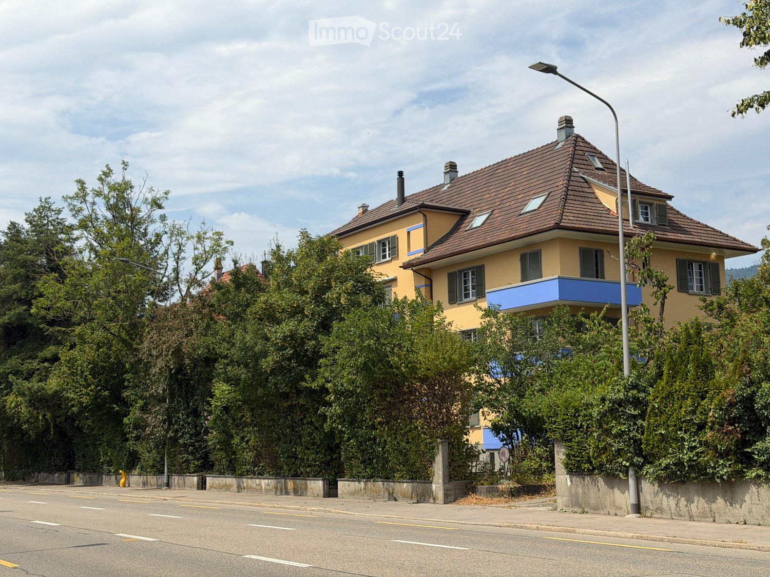 Two-story yellow house with brown tiled roof, multiple windows, and a blue balcony on the second floor, surrounded by greenery and trees.