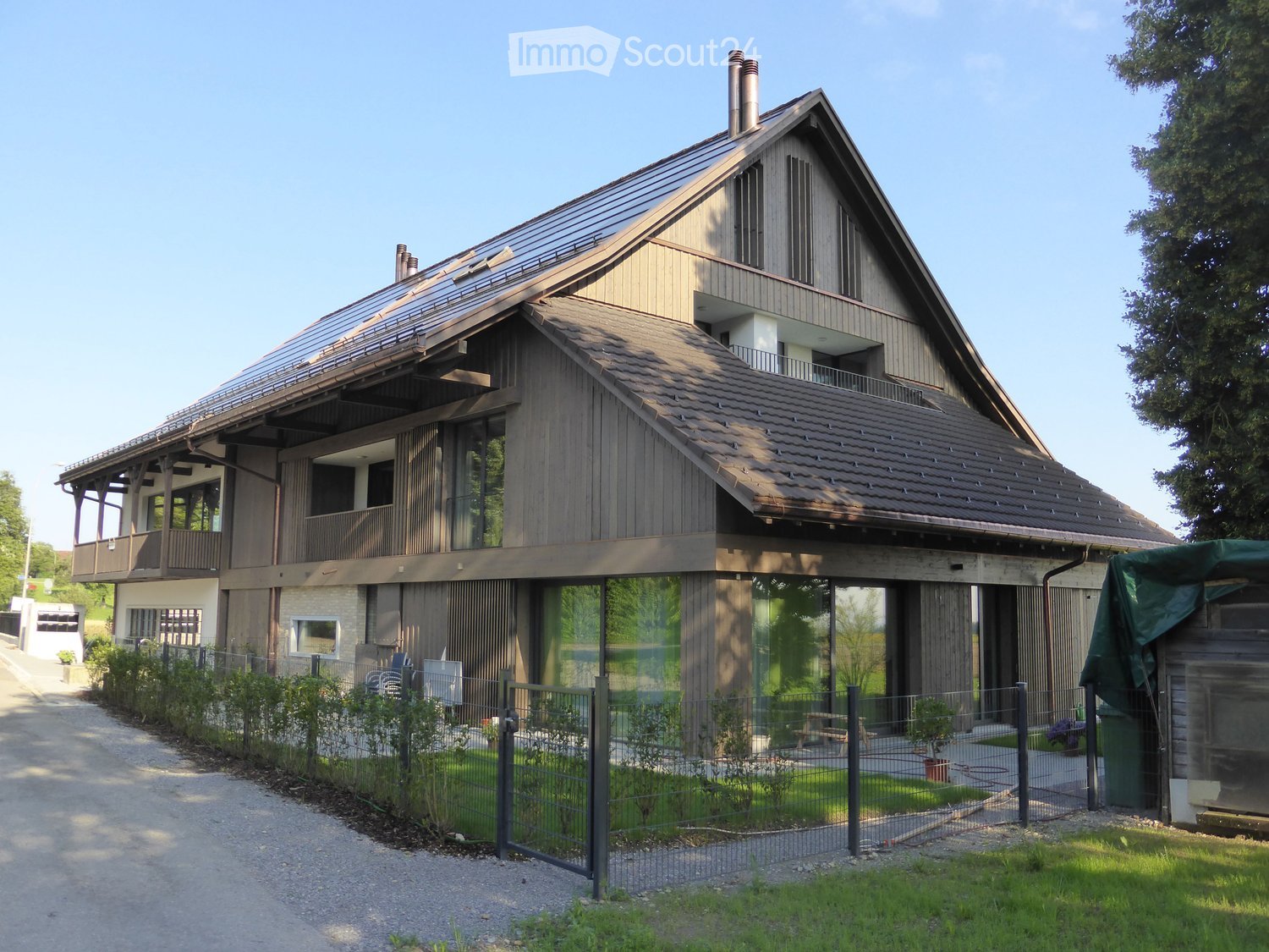 Two-story modern house with wooden exterior, large windows, and a balcony on the upper level. The house features a sloped roof and is surrounded by a well-maintained garden with a gravel driveway.