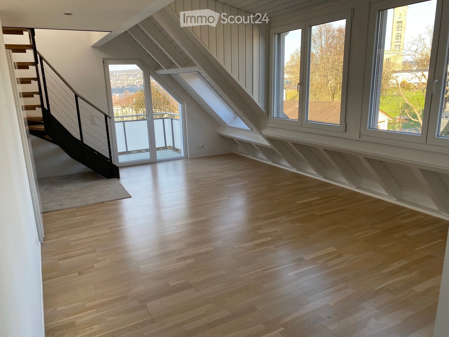 Empty room with wooden flooring, white walls, large windows, and a staircase leading upwards.