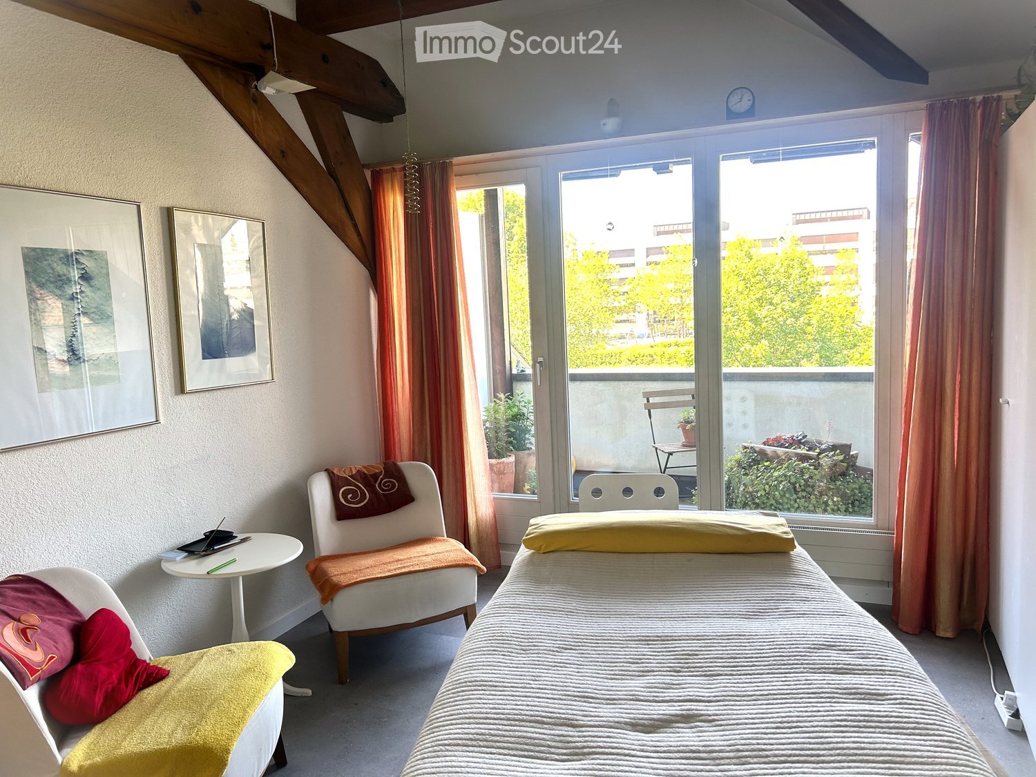 Cozy bedroom with a single bed, two chairs, a small round table, and framed pictures on the wall. Large windows with curtains lead to a balcony.