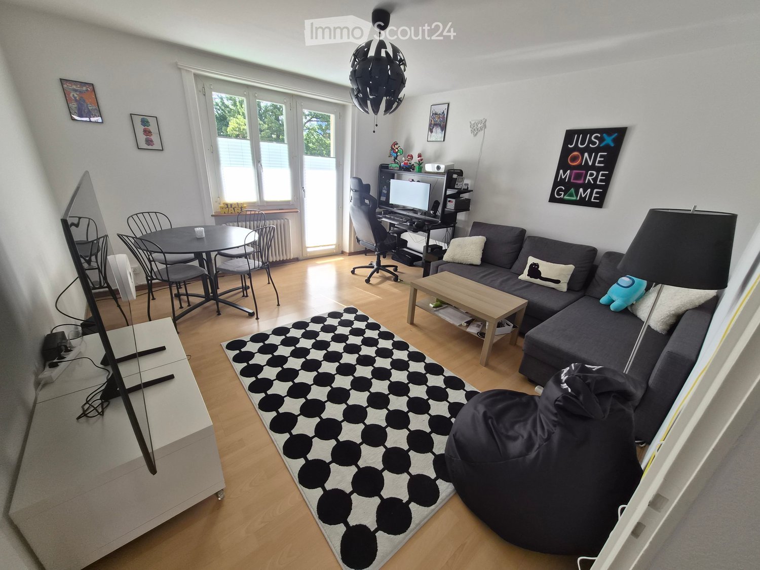 A modern living room with a grey sofa, coffee table, dining table, chairs, TV screen, and a hanging lamp. The room features a black and white patterned rug and a large window with a view of greenery.
