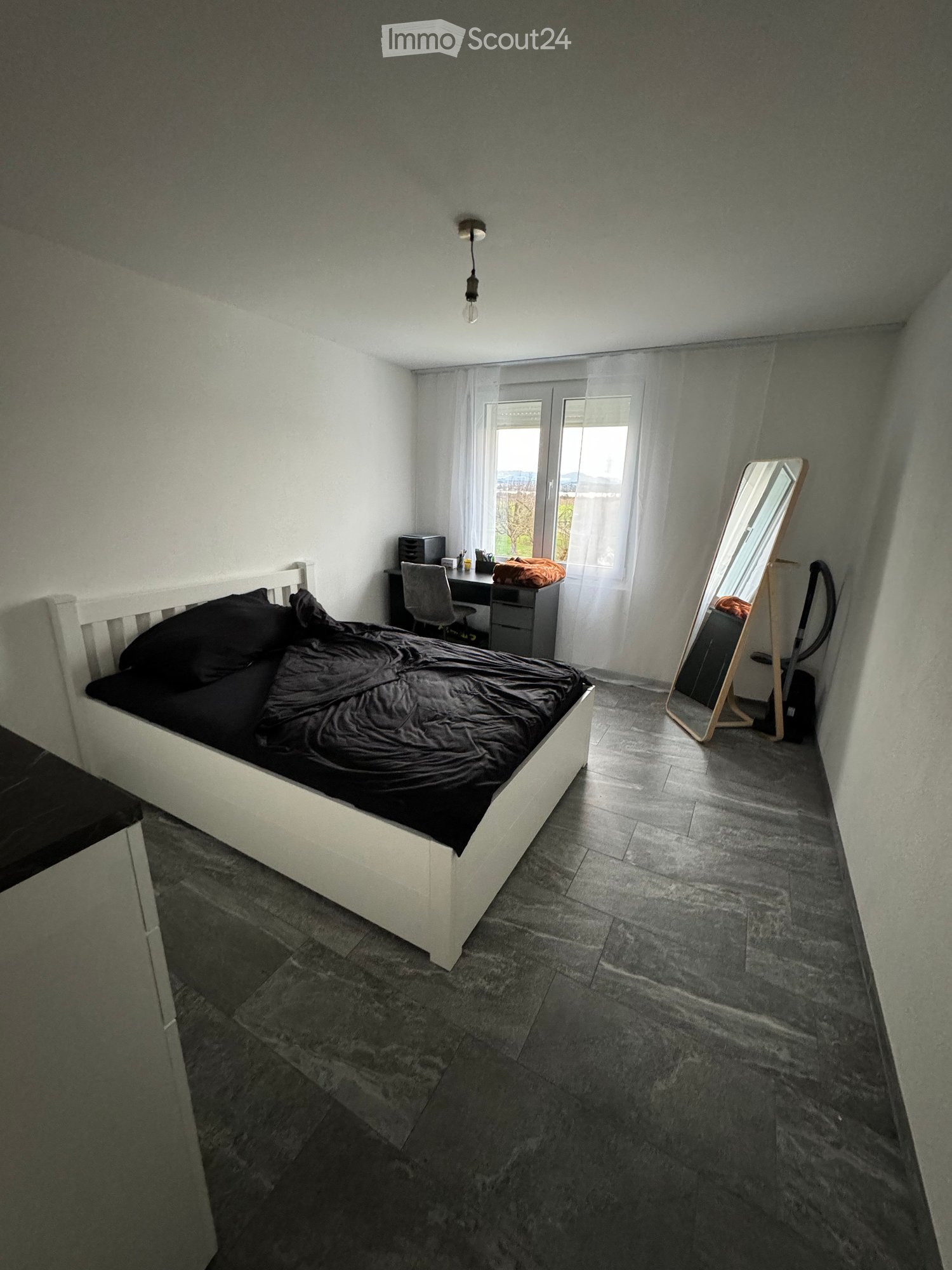 Modern bedroom with a white bed, dark bedding, a large mirror, a desk with a chair, and a lamp hanging from the ceiling.