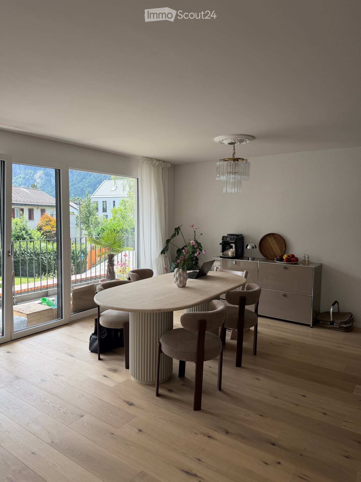 Modern kitchen with wooden floor, large round table, six chairs, laptop, vase, coffee machine, cabinet, and a view of the outdoor area.
