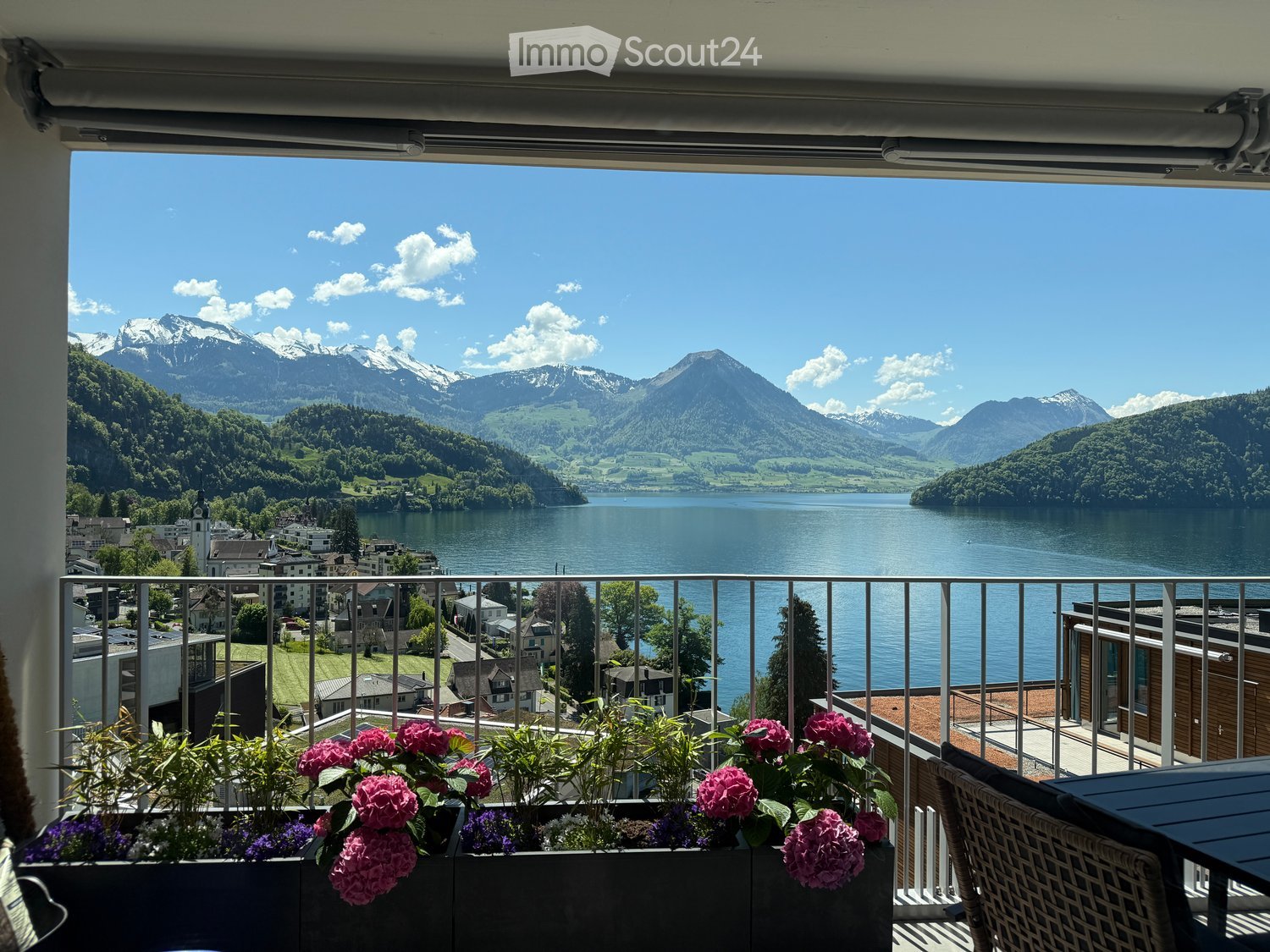 Balcony with a scenic mountain and lake view, decorated with pink flowers in planters, and a small seating area with a table and chair.