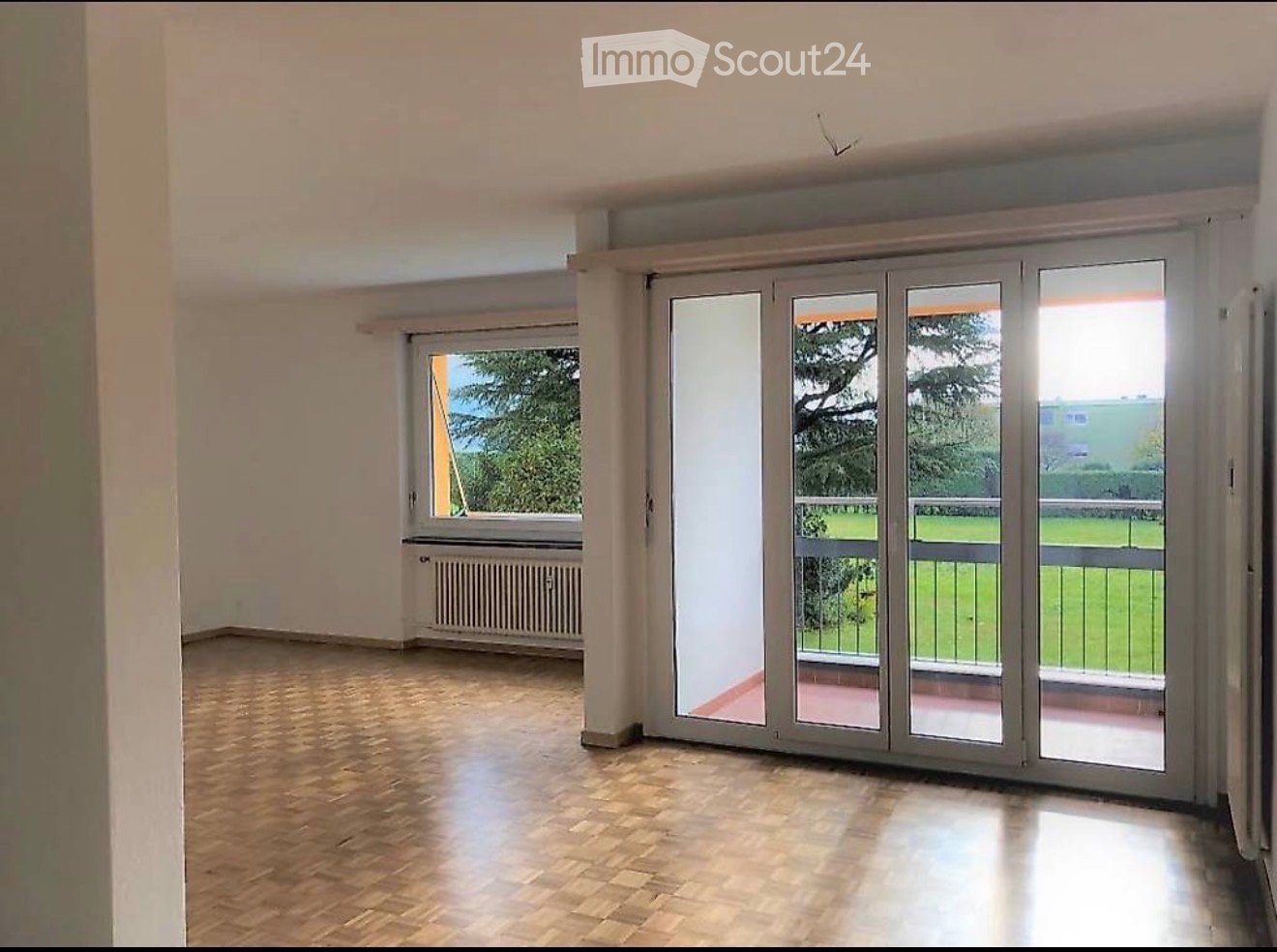 Empty room with wooden flooring, large sliding glass doors leading to a balcony, and a window with a view of greenery.