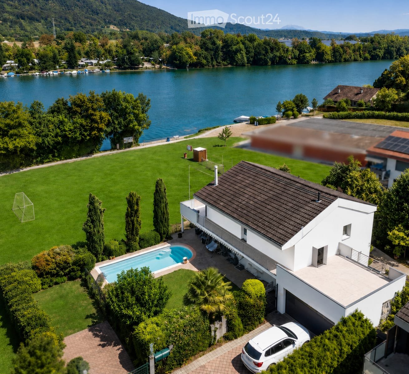 A modern two-story house with a dark roof and white walls, situated near a lake with a swimming pool, lawn, and surrounding greenery.