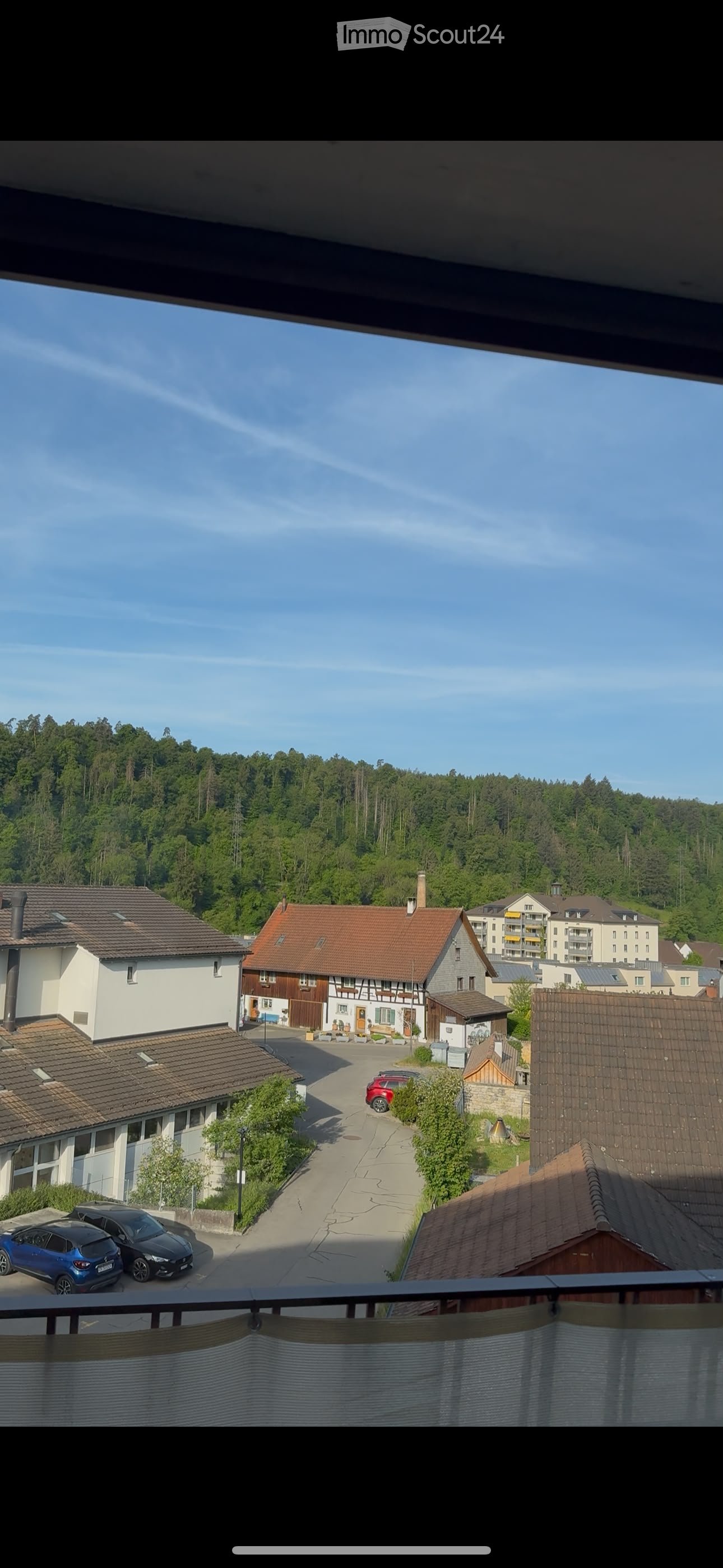 View from a balcony overlooking a quiet residential street with houses, parked cars, and a forested hill in the background.