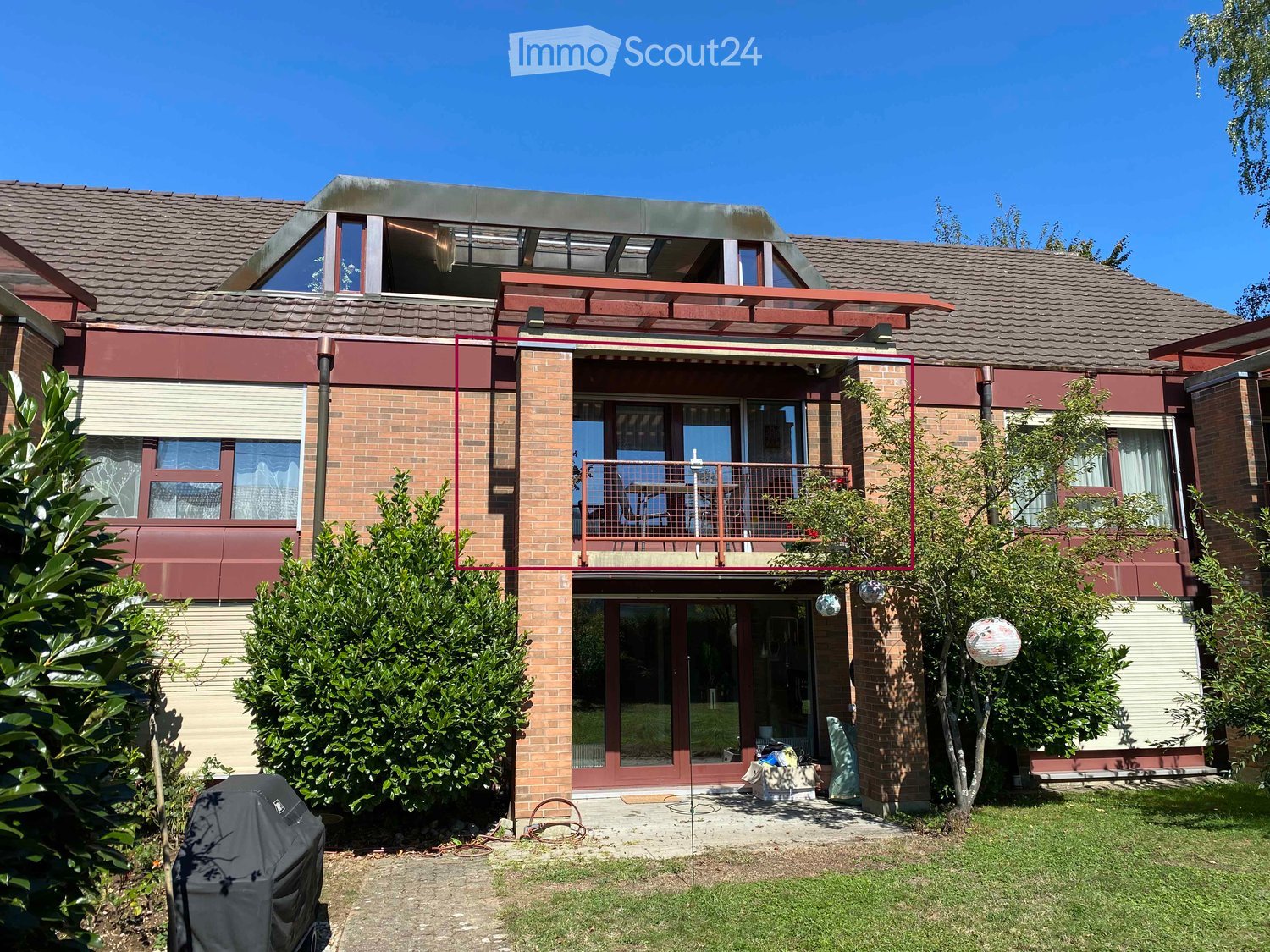 Two-story brick house with a modern design, featuring a large balcony with a railing, multiple windows, and a well-maintained garden with shrubs and a covered grill.