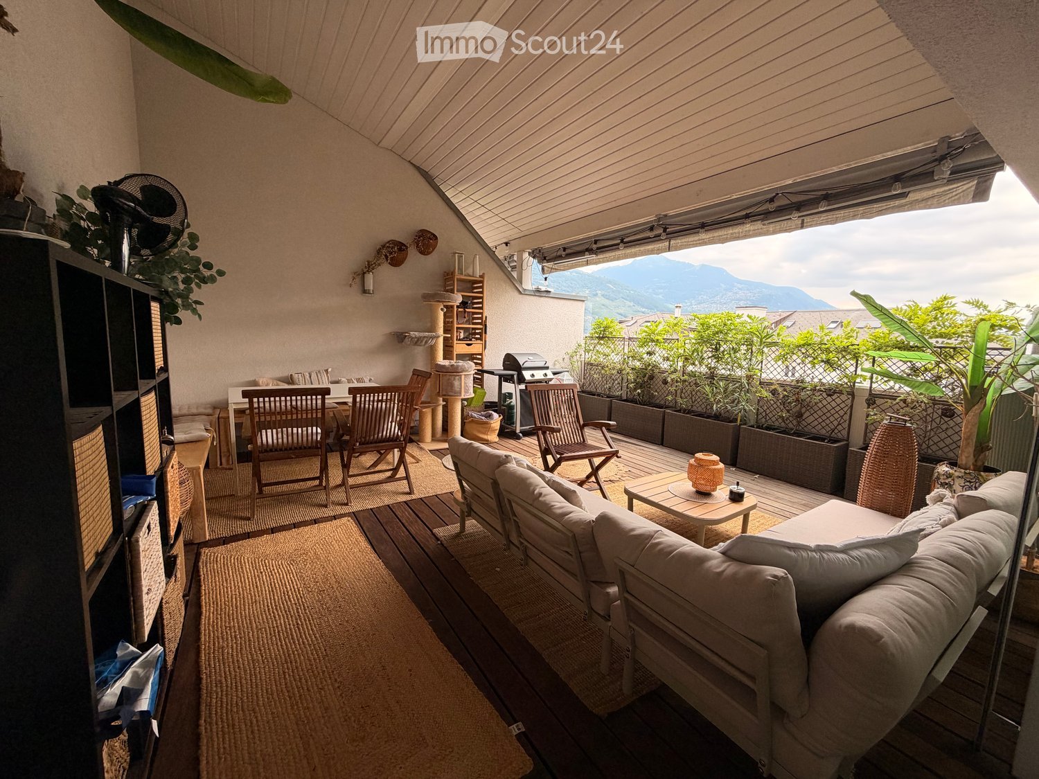 A furnished balcony with a sofa, coffee table, chairs, a fan, cat tree, and plants, offering a scenic mountain view.