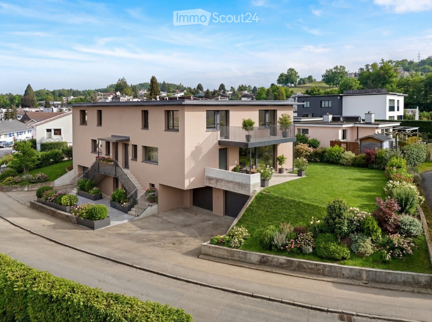 Modern two-story house with a raised ground floor, garage, and balcony. The house features a light pink facade, large windows, and a well-maintained garden with various plants and shrubs.
