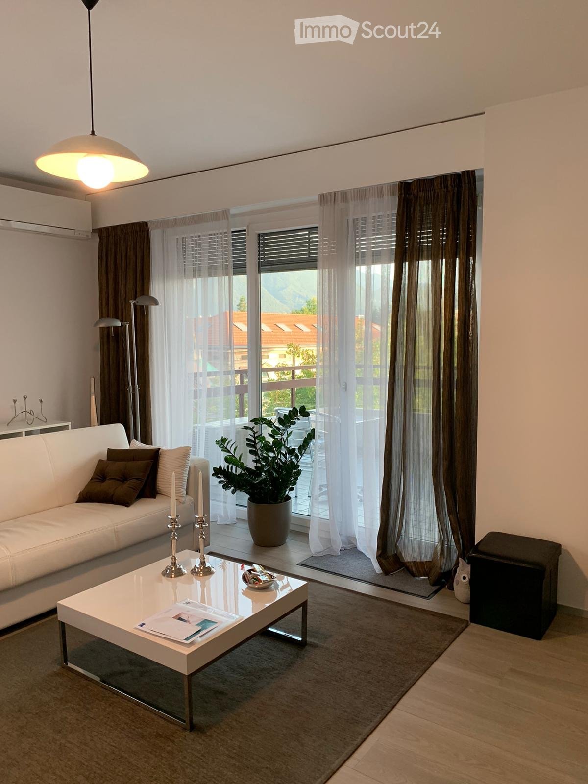 Modern living room with white sofa, coffee table, candles, and a potted plant near large windows with sheer curtains.