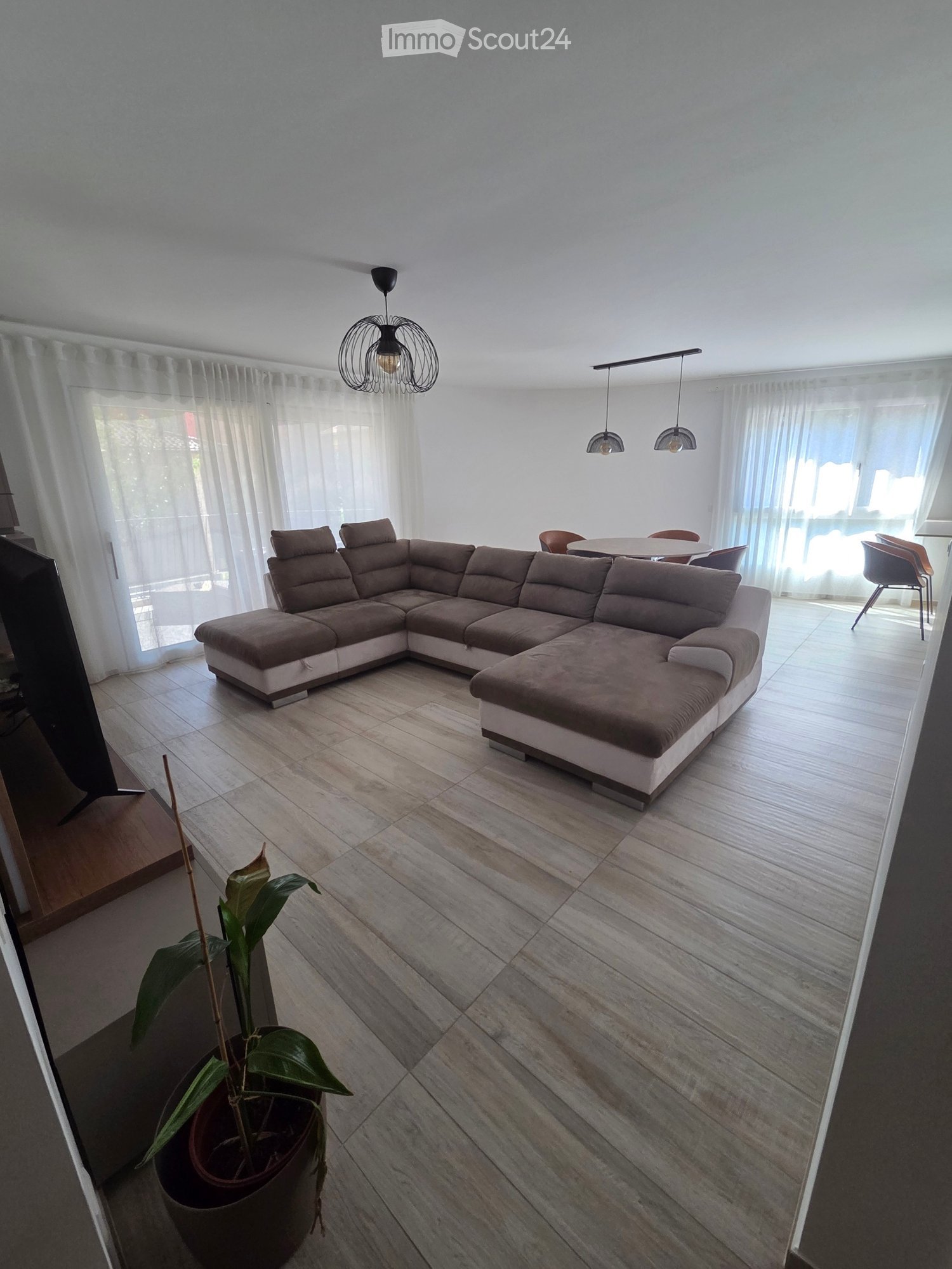 Modern living room with a large L-shaped sofa, a dining area with a round table and chairs, pendant lights, and a potted plant near the entrance.