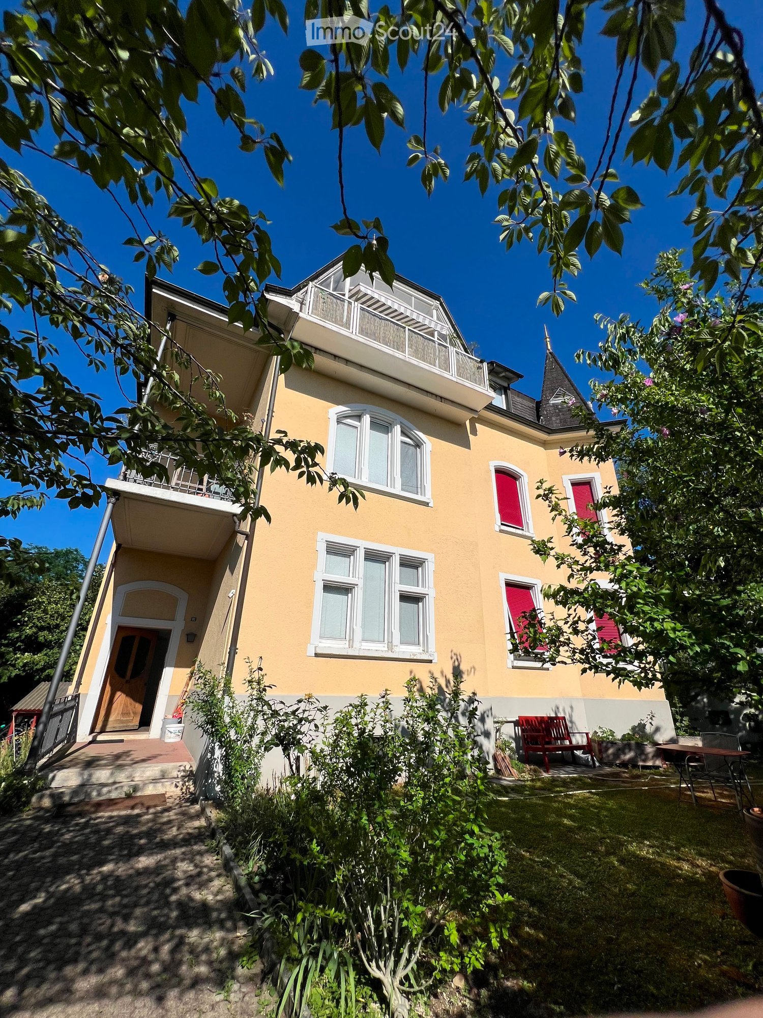 Two-story yellow house with white-framed windows, red shutters, and a small balcony on the upper floor. The house has a well-maintained garden with a bench and a table.
