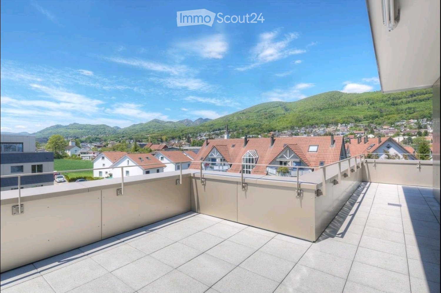 Balcony with tiled floor, low beige walls, metal railing, and scenic view of town and mountains.