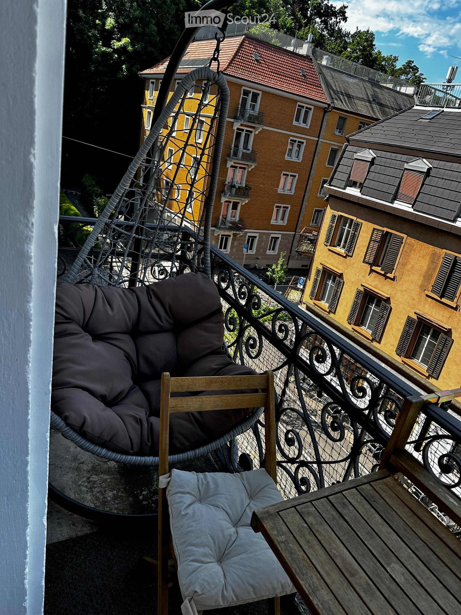 Balcony with a wooden table, a cushioned chair, and a hanging wicker chair, overlooking a cityscape with yellow buildings and balconies.
