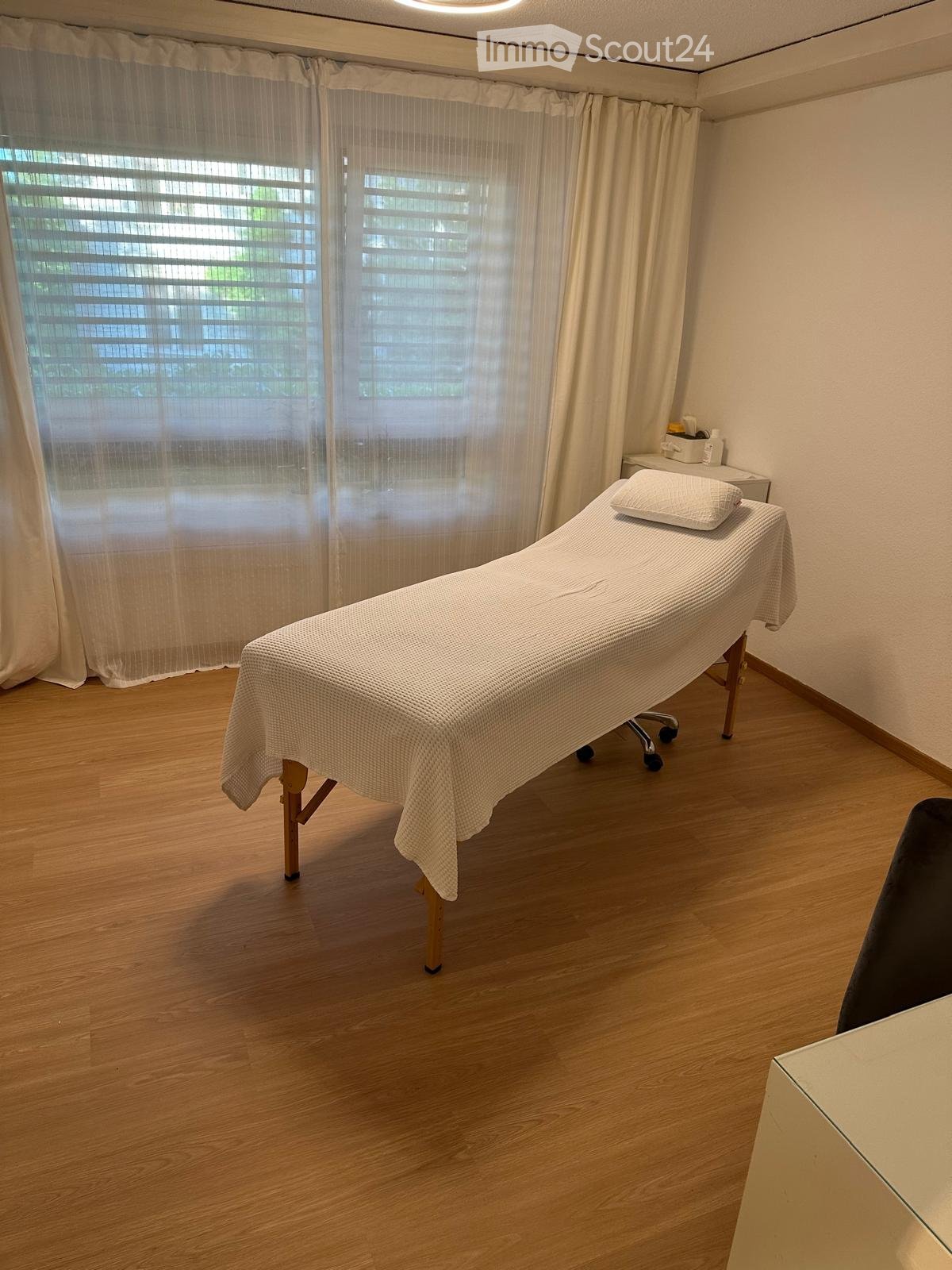 A clean, well-lit room with a wooden floor, a massage table covered in white linens, a small cabinet, and large windows with blinds.