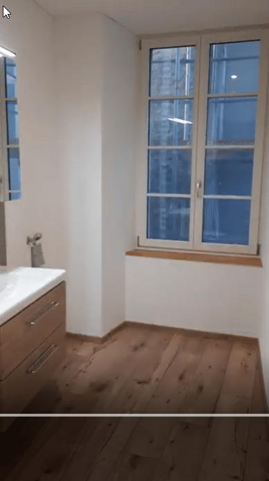 Empty room with wooden flooring, white walls, and large windows. Minimalist design with a small cabinet and sink visible.
