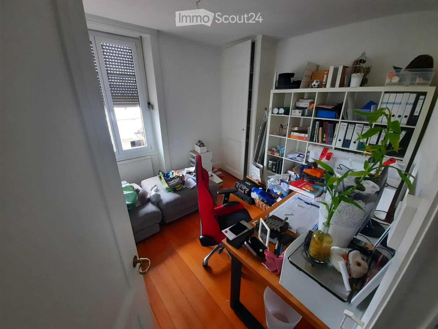 A furnished room with a wooden floor, a red office chair, a desk with a laptop and screen, a sofa, a bookshelf, and a plant on the desk.