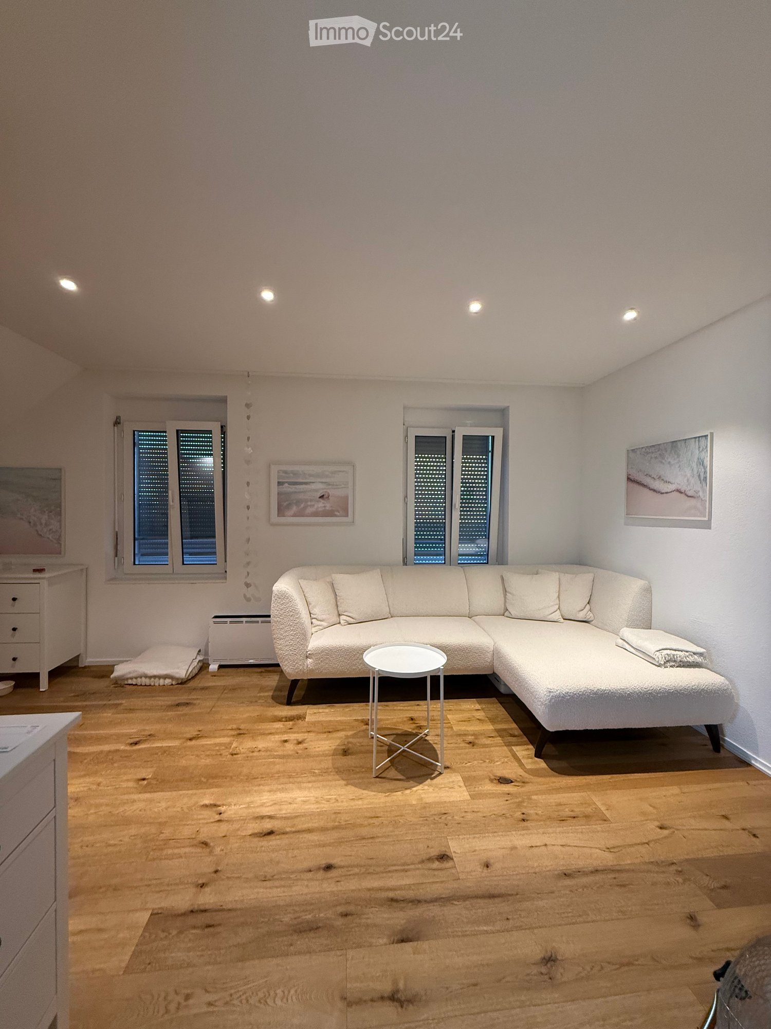 Modern living room with light wooden flooring, white walls, and large windows. Features a white sectional sofa, a small round coffee table, a side table, and picture frames on the walls.