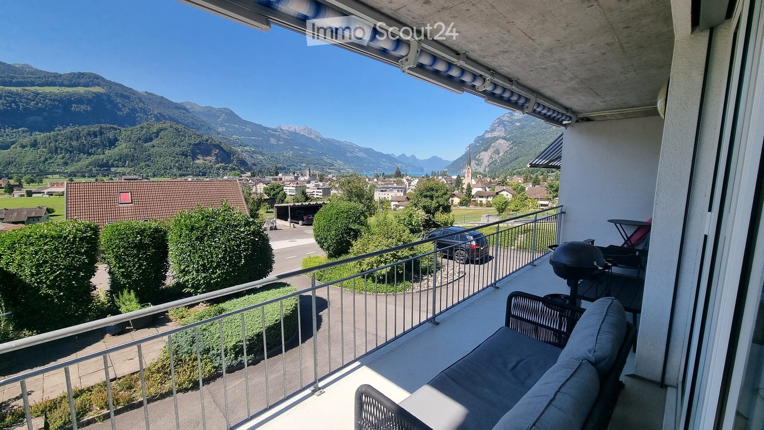 Balcony with a view of mountains and town, equipped with a sofa, chairs, a table, and a grill.