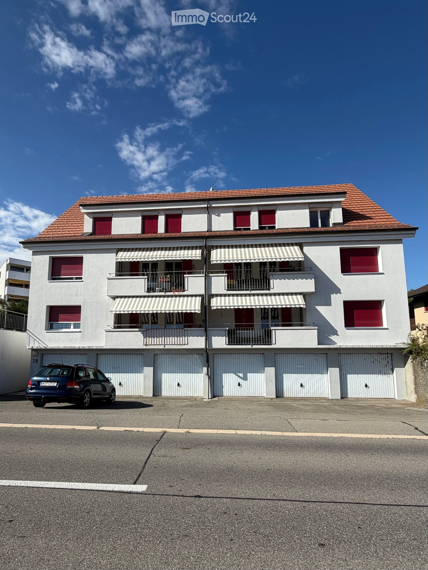 A two-story house with white walls, red shutters, and a red-tiled roof. Features include multiple balconies with striped awnings, a garage area with closed garage doors, and a parked car in front.
