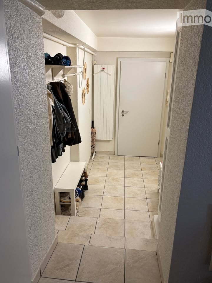 A narrow hallway with tiled flooring, white walls, and built-in storage shelves holding shoes and coats. A closed white door is visible at the end of the hallway.