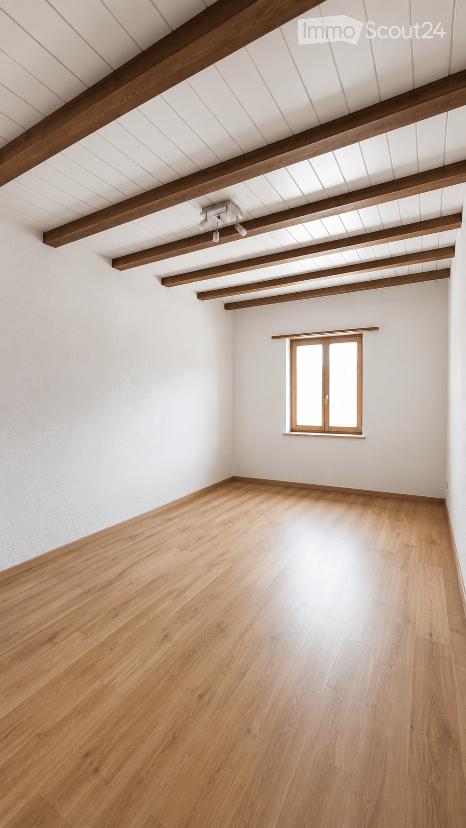 Empty room with wooden flooring, white walls, exposed wooden beams on the ceiling, and a single window with wooden frame.