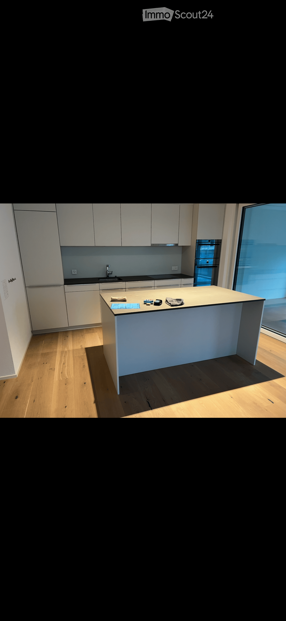 Modern kitchen with white cabinets, a central island, and a sink area. The floor is wooden, and the space is well-lit with natural light from a large window.
