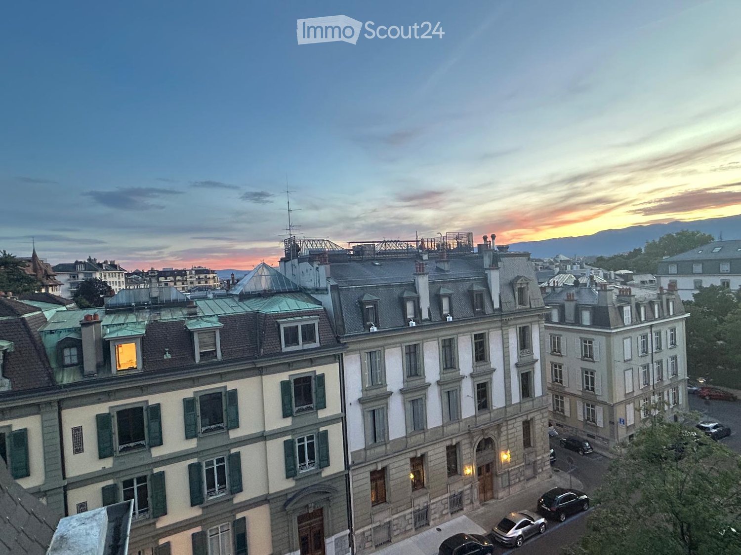 A panoramic view of a cityscape at sunset, featuring multiple multi-story buildings with varied architectural styles, including green shutters and stone facades, with visible rooftops and chimneys.