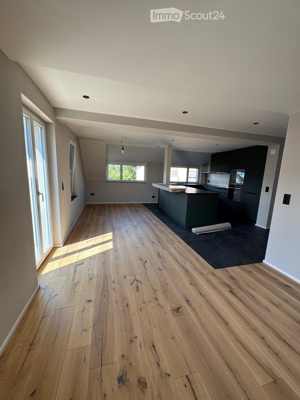 Empty room with light wooden flooring, large windows, and a view of the kitchen area with dark cabinets and a countertop.