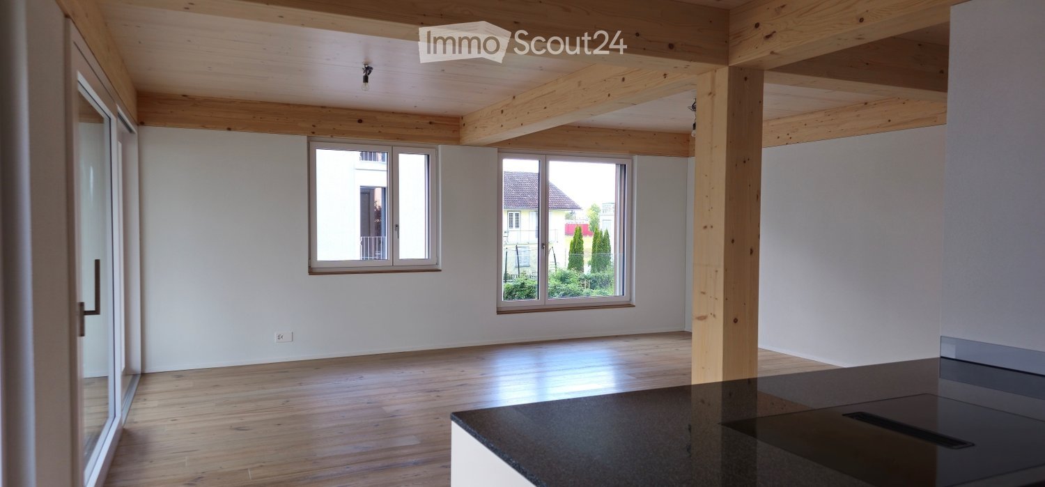 Bright, open room with wooden beams on the ceiling, large windows, and a polished wooden floor. The space is completely empty, with no furniture or decorations.