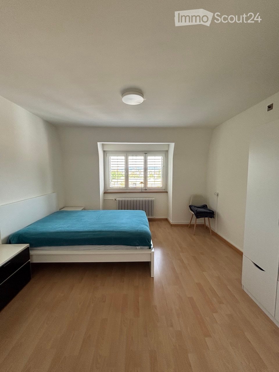 A furnished bedroom with a wooden floor, a single bed with blue bedding, a nightstand, a chair, and a radiator beneath a window.