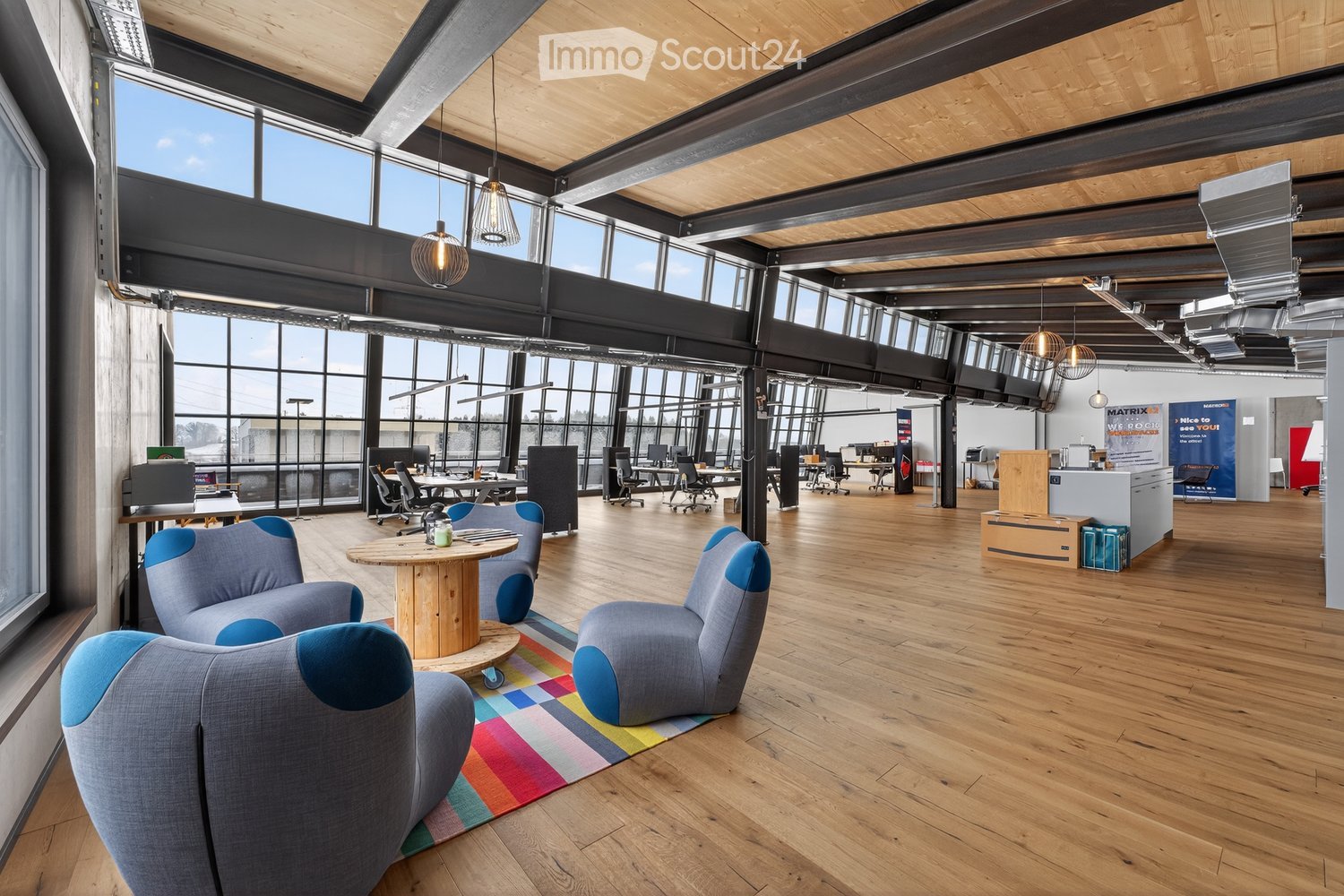 Modern office space with wooden floors, large windows, industrial-style lighting, and seating area with a round wooden table and blue chairs.