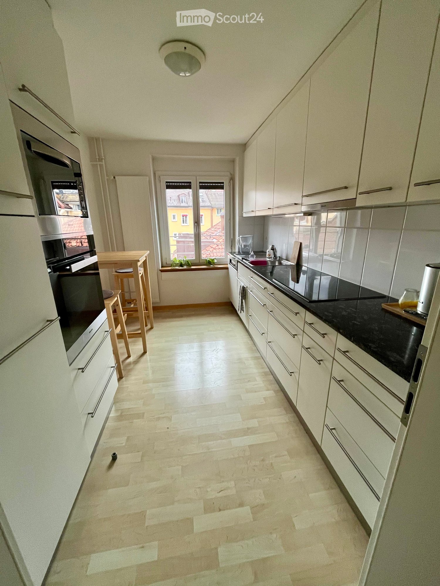 Modern kitchen with white cabinets, black countertops, and a central island. Features include a sink, oven, microwave, and a small dining area with stools.