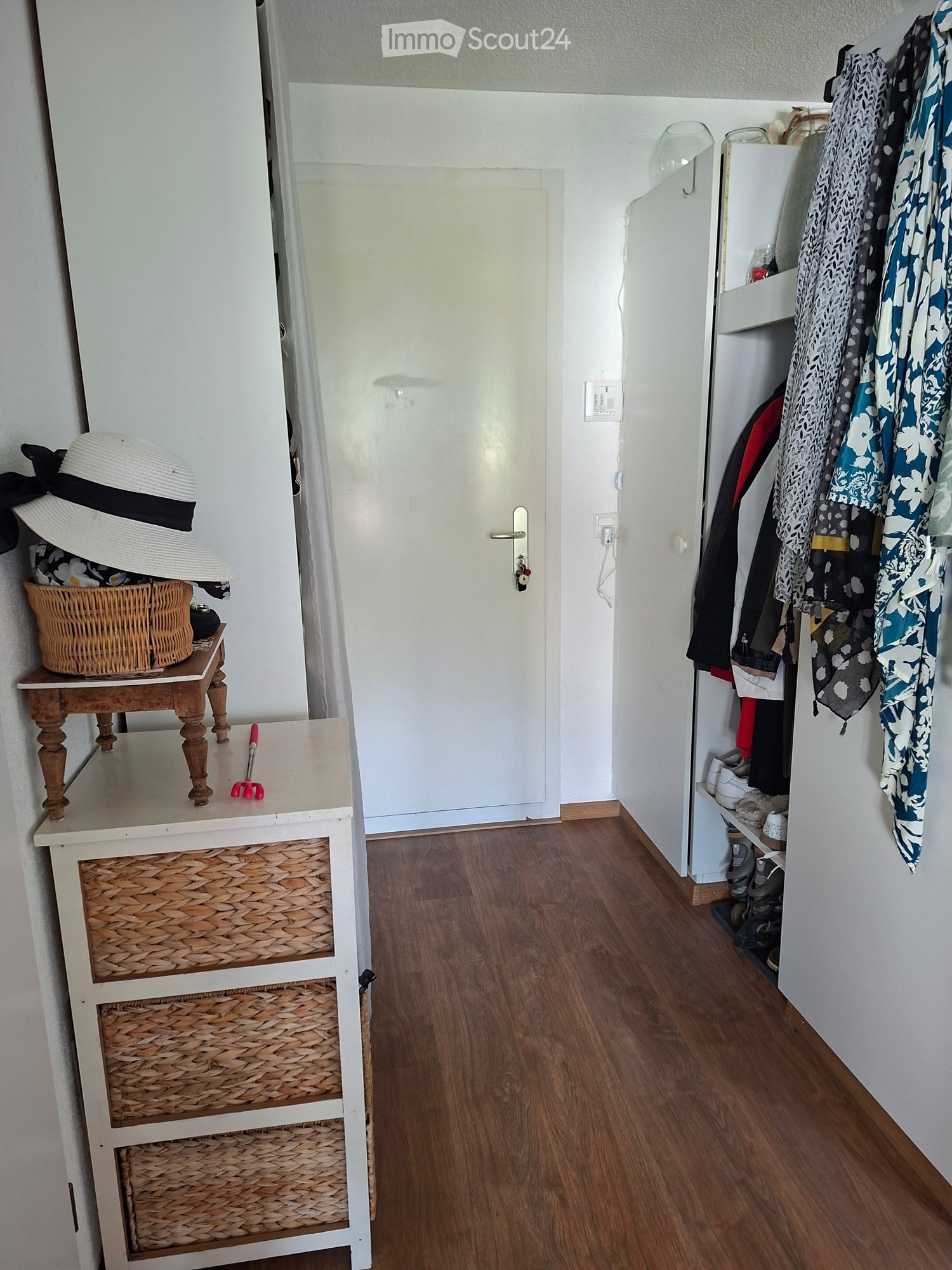 Hallway with wooden flooring, white walls, a small wooden table with a woven basket and hat, a coat rack with clothes, and a door leading to another room.