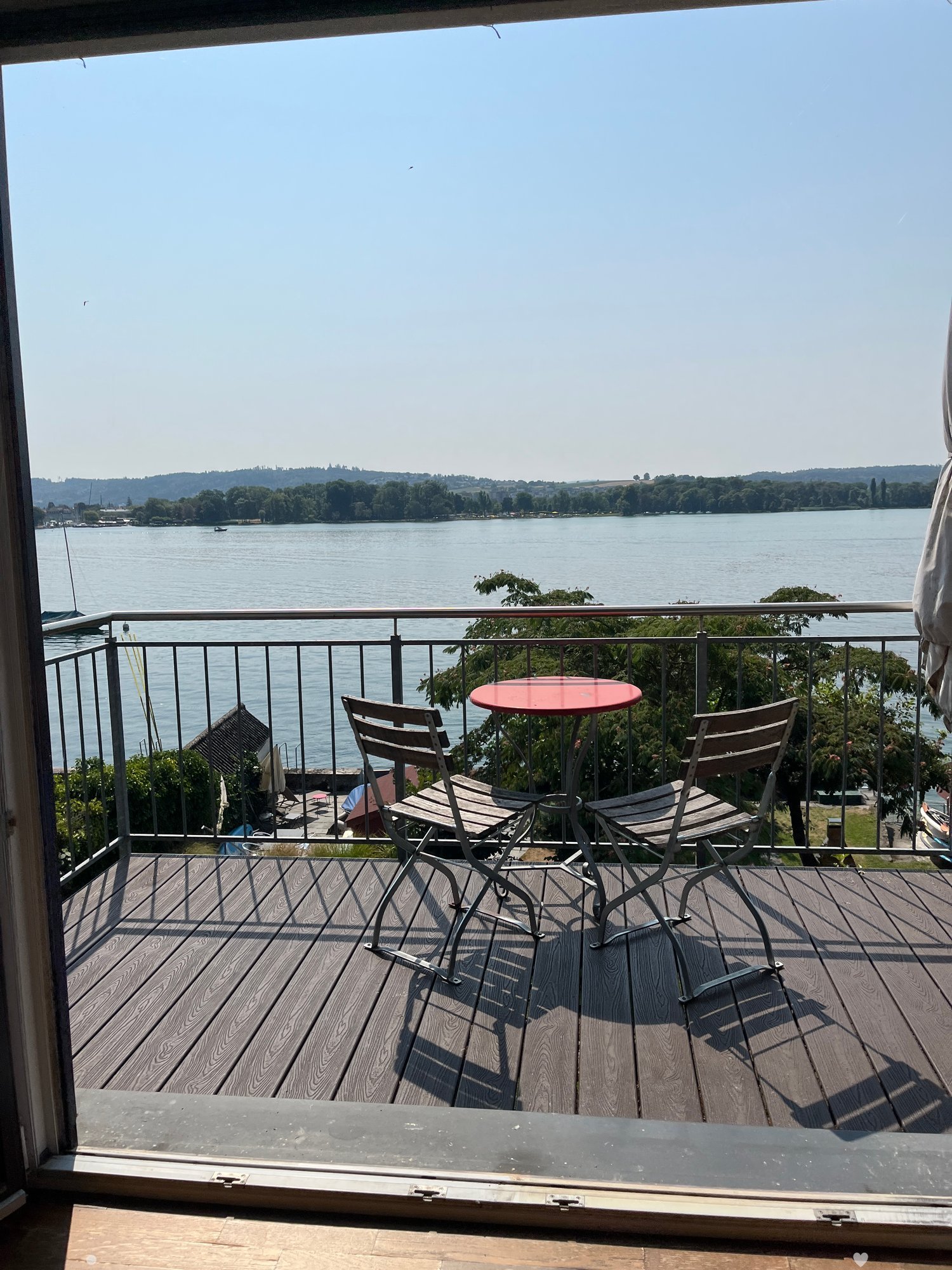 Balcony with wooden decking, metal railing, and a red round table with two wooden chairs, overlooking a lake.