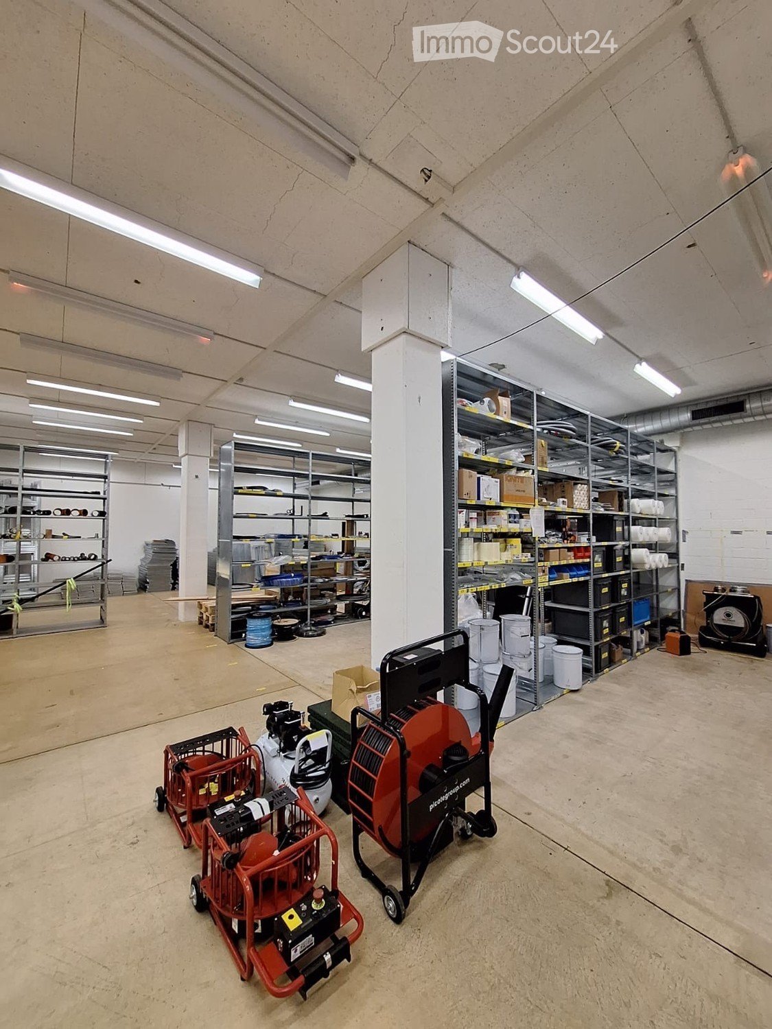 Empty warehouse space with concrete floors, metal shelving units, and industrial equipment like generators and air compressors.