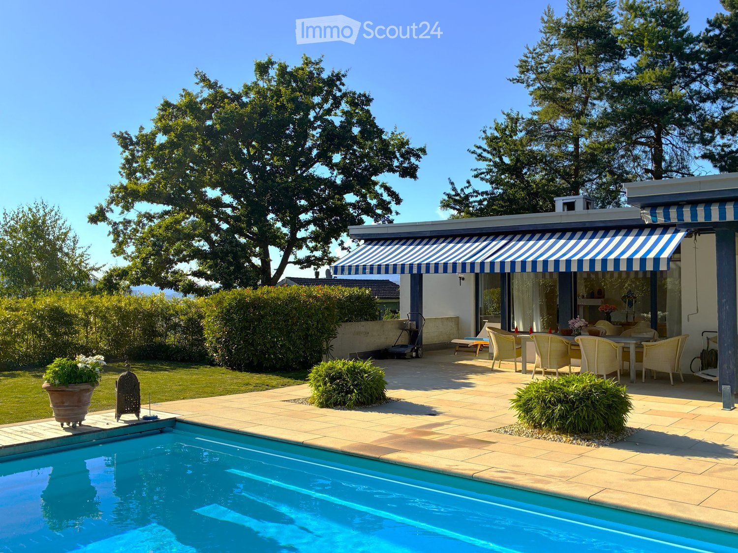 Outdoor patio area with a swimming pool, lounge chairs, dining table, and surrounding greenery.