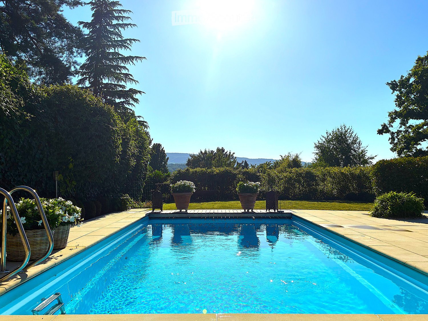 A rectangular swimming pool surrounded by a paved deck, with potted plants and lounge chairs nearby. The background features a well-maintained lawn, trees, and a clear blue sky.