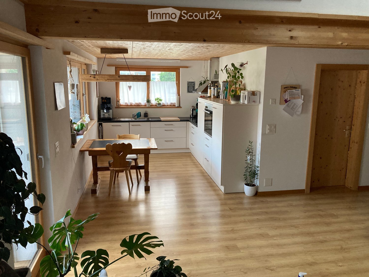 Bright, open kitchen with wooden floors, white cabinets, a central dining table with chairs, and various appliances including a refrigerator, oven, and coffee maker. Natural light streams in from large windows, complemented by potted plants.