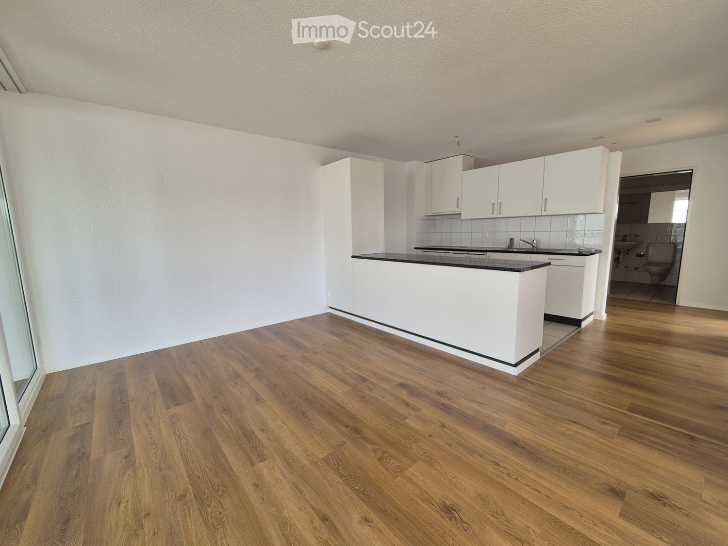 Empty kitchen with white cabinets, black countertop, and wooden flooring. Adjacent bathroom visible with white tiles and fixtures.