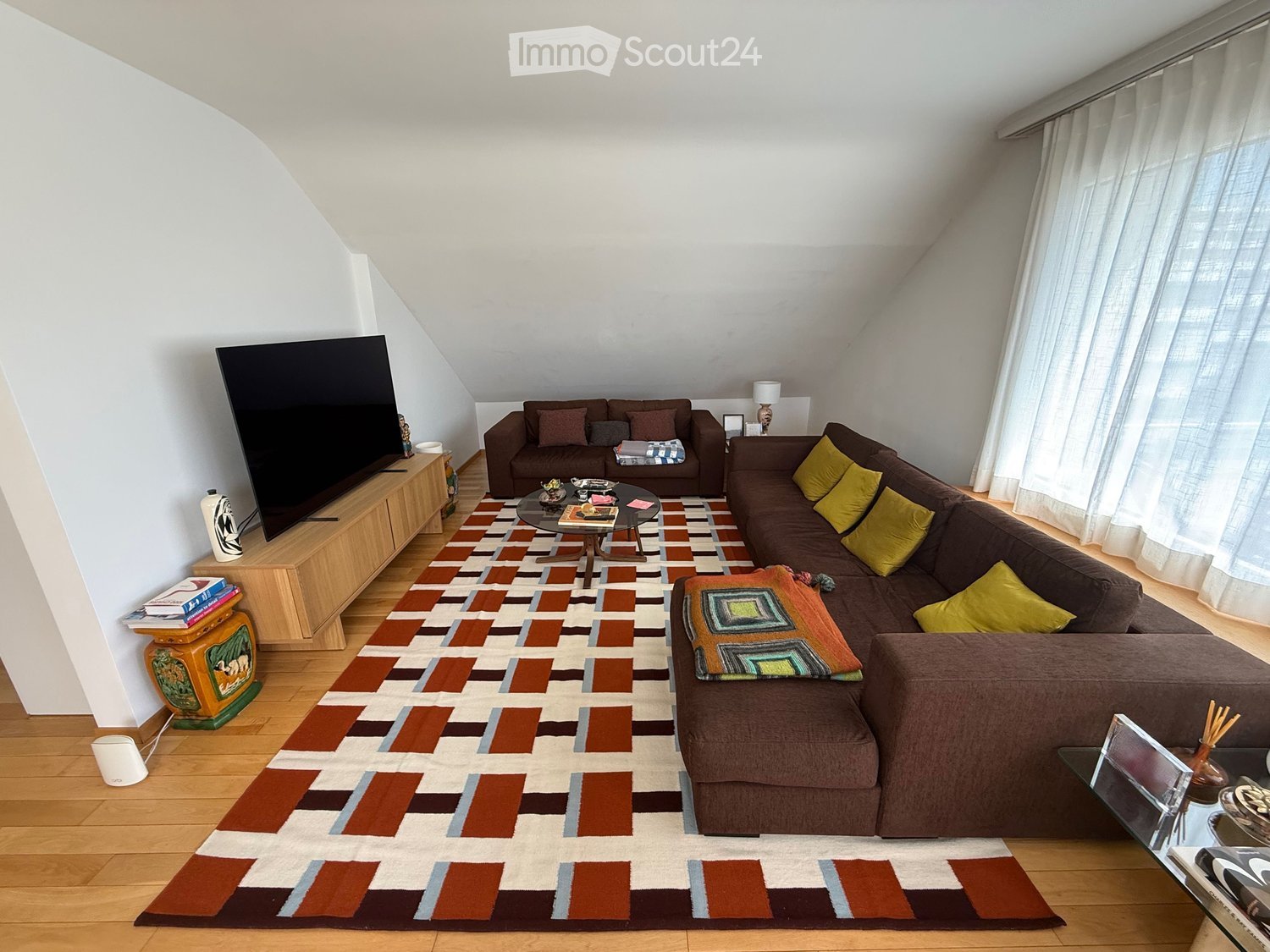 Living room with wooden flooring, large window with white curtains, brown sectional sofa, coffee table, TV, and patterned rug.